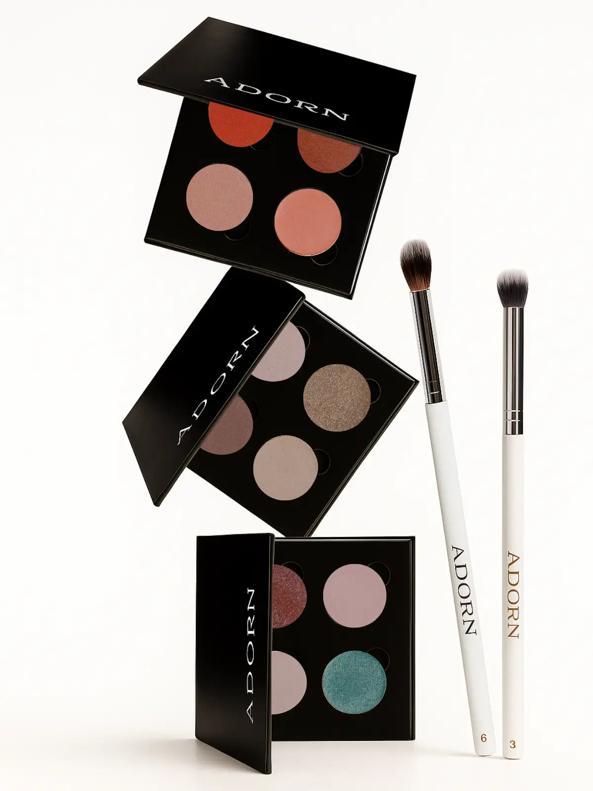 Makeup product set with three eyeshadow palettes and two brushes on a white background, branded 'Adorn'.