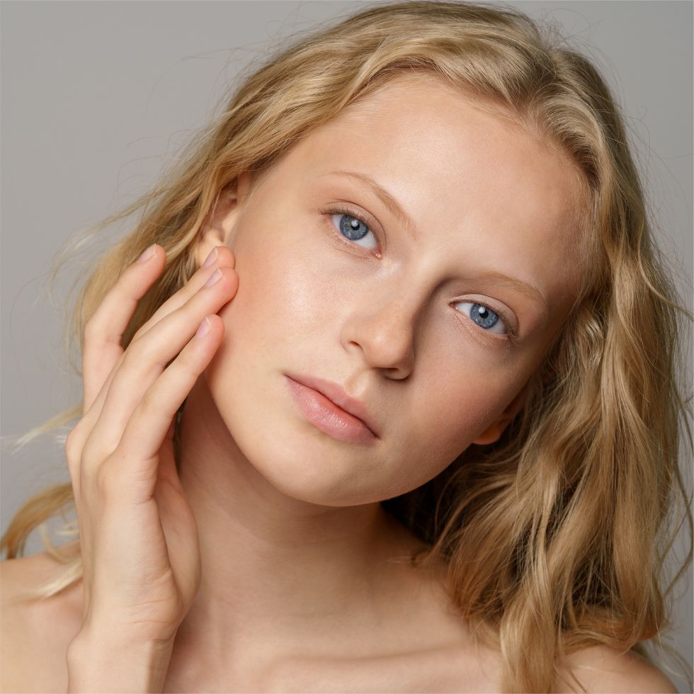 Woman with blue eyes and blonde hair touching her face against a neutral background