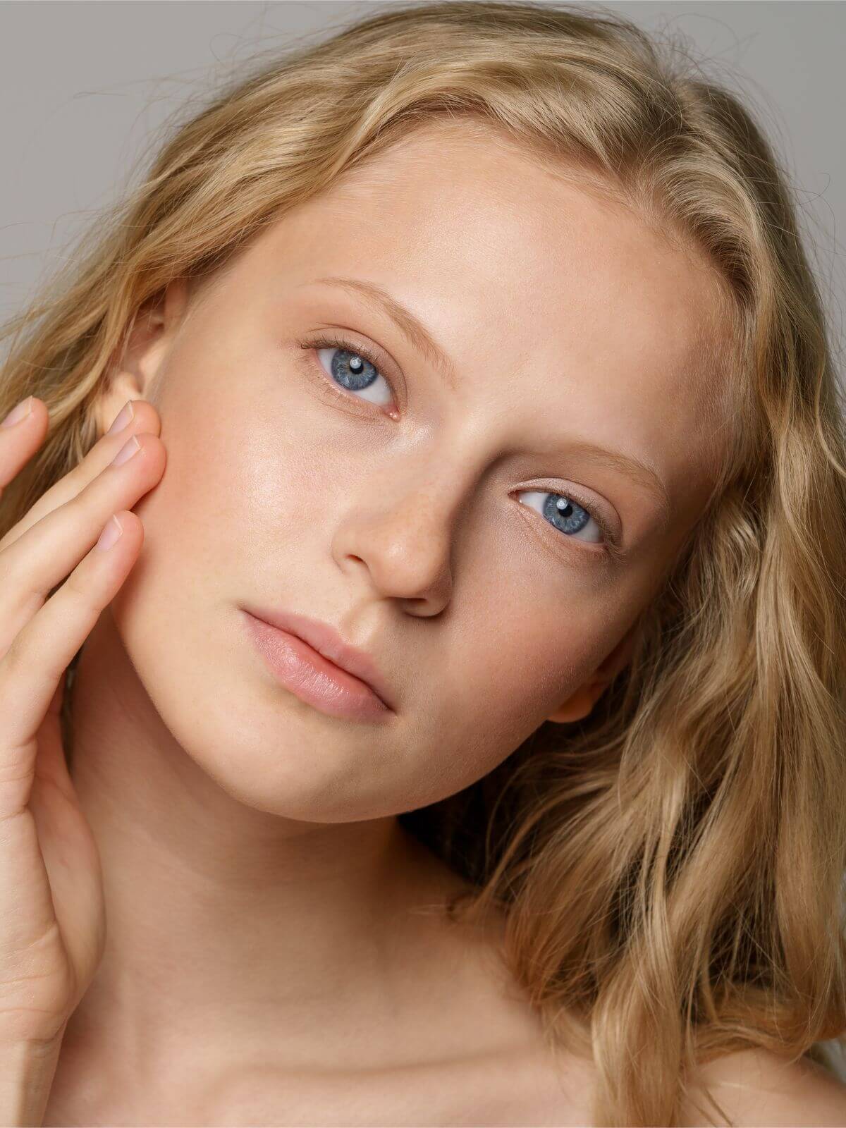 Close-up of a person with blonde hair and blue eyes, touching their cheek.