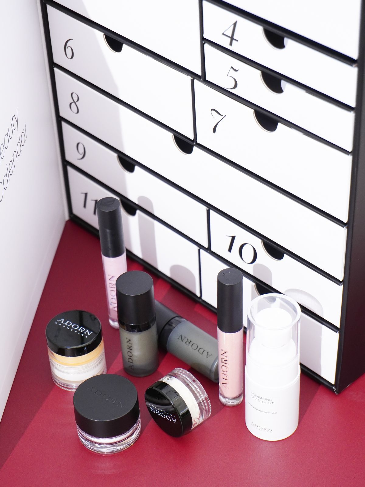 Makeup products in front of a black and white advent calendar beauty numbered drawer set on a burgundy surface.