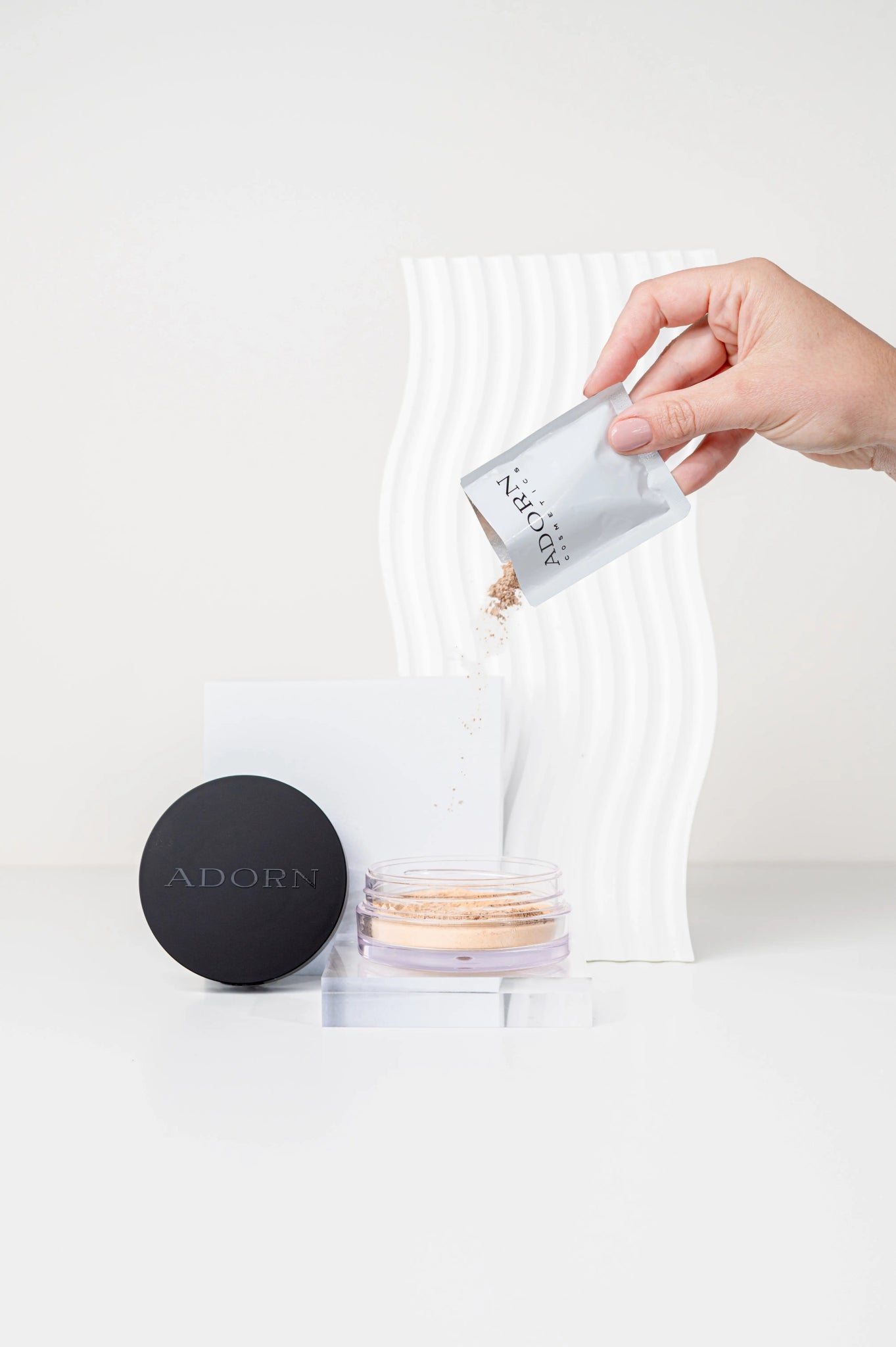 Hand pouring mineral foundation powder into a container with 'ADORN' branding on a white background.